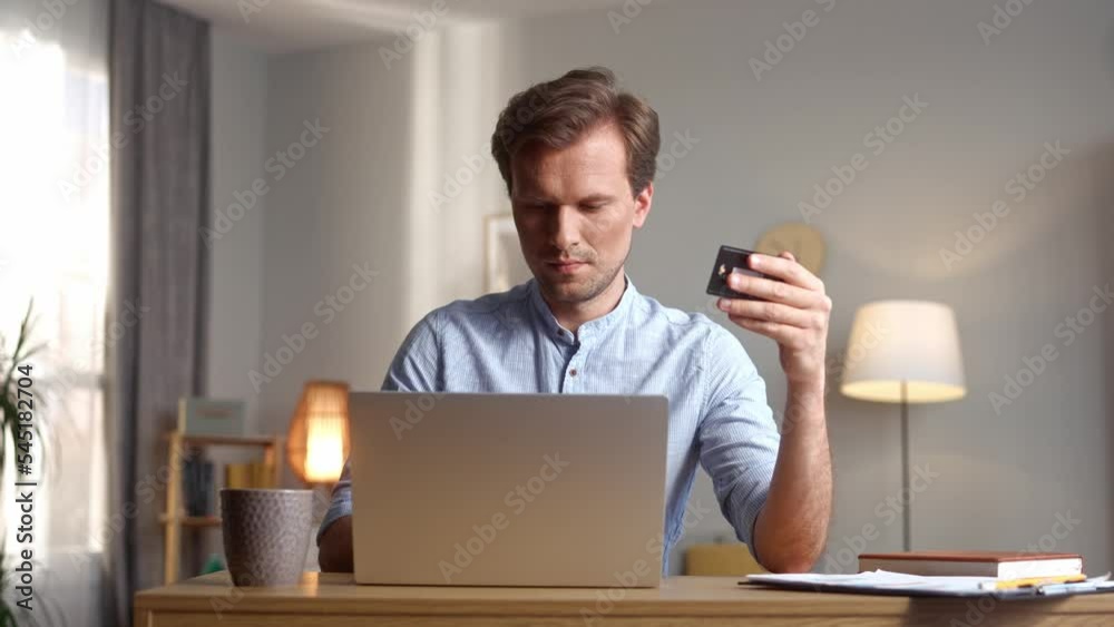 Successful young elegant male making payments online using laptop holding credit card. Happy attractive man shopping on Internet buying things sitting at home in cosy living room.