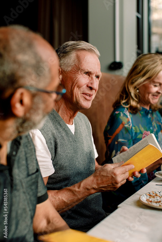 Senior man holding book while talking with male and female friends in cafe photo