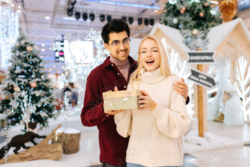 Portrait of happy amazed young woman receiving gift box from loving boyfriend standing in hall of celebrate shopping mall in Christmas eve, on background of bright beautiful xmas decorations.