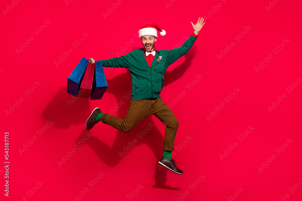 Full length photo of excited charming guy dressed xmas green cardigan jumping rising noel gifts shoppers isolated red color background Full length photo of excited charming guy dressed xmas green cardigan jumping rising noel gifts shoppers isolated red color background