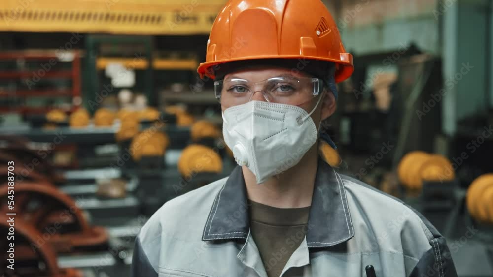 Chest up portrait of confident female plant worker in orange hard hat, safety glasses and face mask looking at camera standing inside of machinery equipment production plant Chest up portrait of confident female plant worker in orange hard hat, safety glasses and face mask looking at camera standing inside of machinery equipment production plant