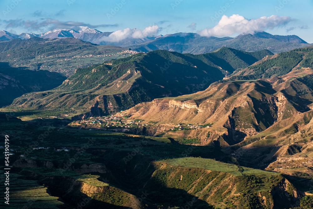 Many high mountains with green landscape and small houses below, clear sunny day.