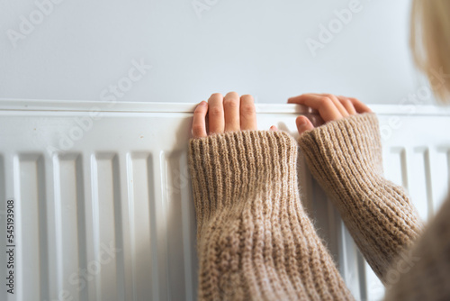 Child wearing brown woolly jacket warming cold hand in front of heating radiator in winter time. Electric or gas heater at home. Heating season, cold room, heating problems