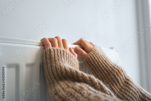 Warming up hands near heater. Young girl in woolly brown sweater warming cold hands in front of heating radiator in cold winter time photo