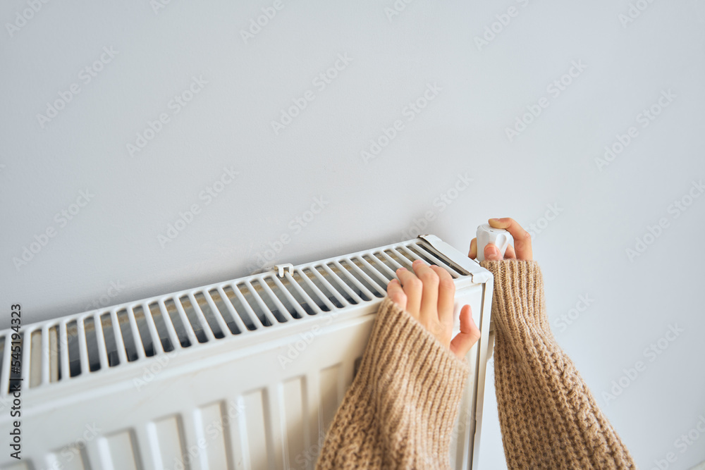 Unrecognized girl sitting near heater radiator at home and adjusting