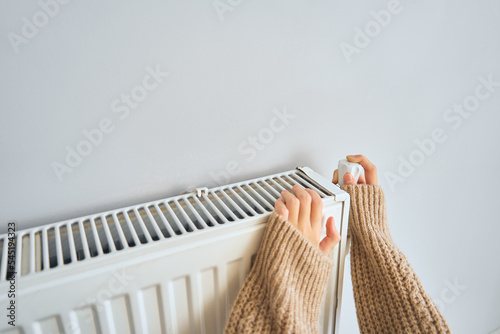 Unrecognized girl sitting near heater radiator at home and adjusting temperature. Electric or gas heater at home. Heating season, cold room, heating problems