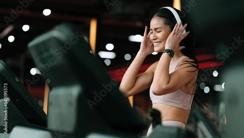 Side view of beautiful young asian woman running on treadmill and listening to music via headphone during sports training in a gym.
