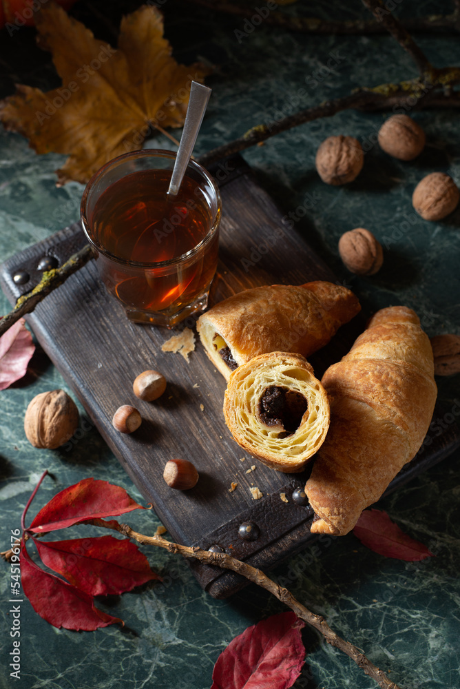 Cozy tea time: a glass on hot tea and chocolate croissants on a wooden board around by pumpkin, autumn leaves, nuts. Close up. Top view