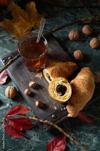 Cozy tea time: a glass on hot tea and chocolate croissants on a wooden board around by pumpkin, autumn leaves, nuts. Close up. Top view