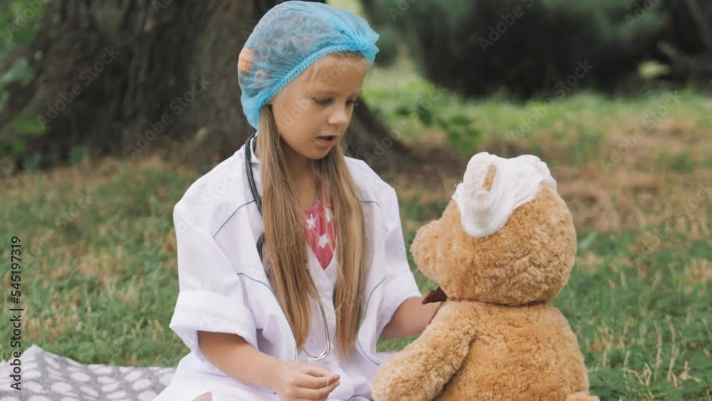 A little girl in a medical white uniform plays with a teddy bear in a hospital in the park.. A little girl in a medical white uniform plays with a teddy bear in a hospital in the park..