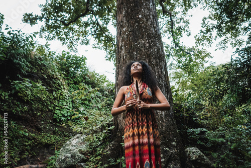 Imagen horizontal desde abajo de una mujer latina de pie en medio de un bosque realizando un ritual mirando hacia el cielo.  photo