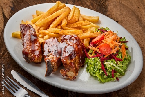 Top view of pork ribs with fries and vegetable salad in a white plate on a wooden table