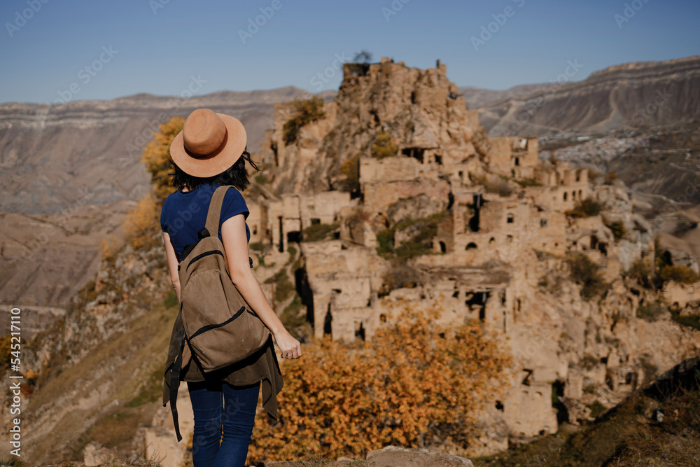 a young tourist girl sits on a wooden bench on the roof of an abandoned house in the ancient village of Gamsutl, Dagestan and enjoys the view of the mountains