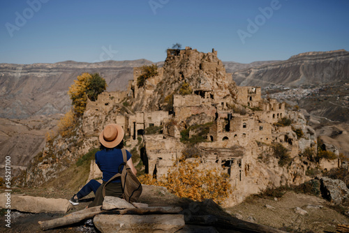 Girl Traveler in a hat with a backpack on a rock looks at the village of Gamsutl
