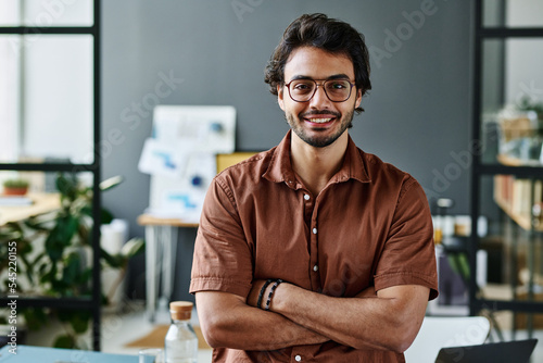 Young successful intern in eyeglasses and brown shirt keeping his arms crossed by chest while standing by workplace in front of camera photo
