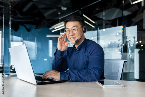 Cheerful and smiling office worker with video call headset, Asian customer support tech of online store, smiling and talking consulting. the man works inside the office.