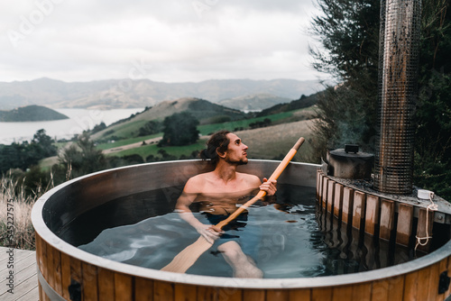 calm caucasian boy without clothes taking a bath with a long wooden spade in his hands inside a metallic hot water jacuzzi relaxing in the mountains far from civilization near the forest trees, te