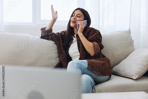 Woman blogger with phone lying at home and talking on the phone on sofa with laptop and working freelancer online, smiling with teeth