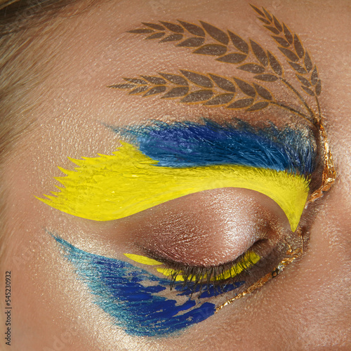Art makeup on woman eye. Large photo frame. Square composition. Ukrainian flag concept, wheats. Golden field of spikelets on skin, face. Civilians against the war in Ukraine. Stop Russian aggression