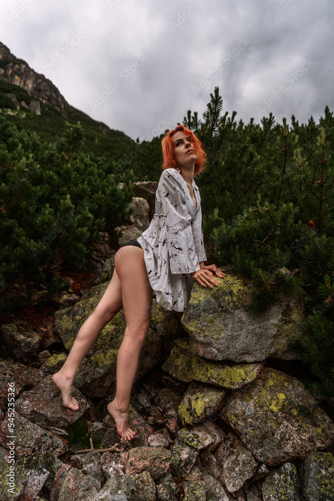 Elegant sexy woman with red hair in a white dress poses next to rock among spruce in mountains and relaxes under sun rays. Fashion natural portrait as wallpaper, magazine design or advertising cover.