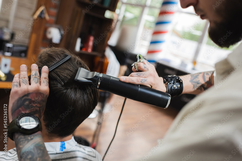 tattooed barber with comb drying hair of client on blurred foreground.