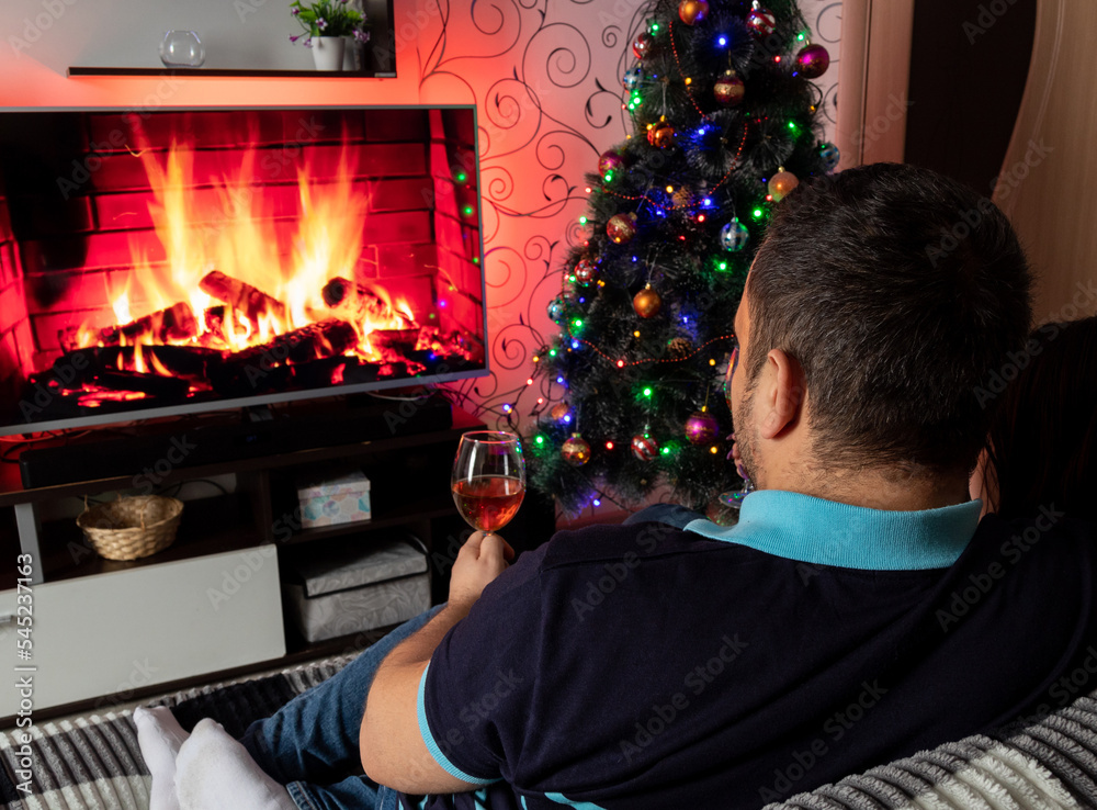 ?ouple is sitting on sofa, holding glass of wine, in front of TV, which depicts fireplace. Selective focus. Images for articles about leisure, winter, family