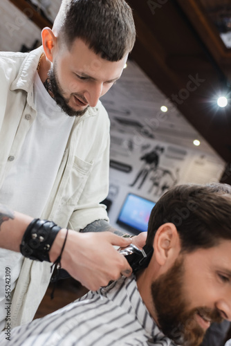 Bearded barber trimming hair of blurred customer in barbershop.