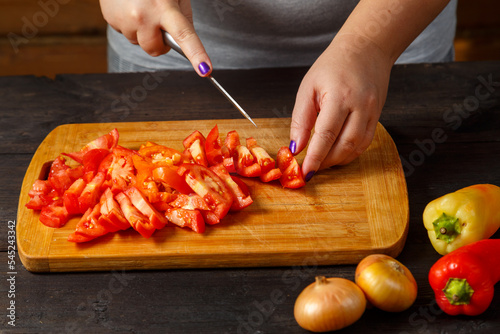 Female hands with a knife cuts red tomatoes on a wooden board on the table next to onions and peppers.