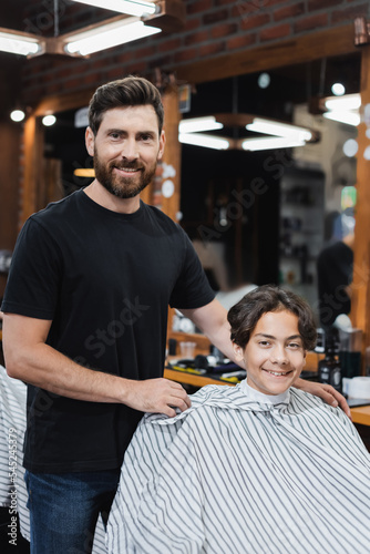 Smiling barber and teenage boy looking at camera in barbershop.