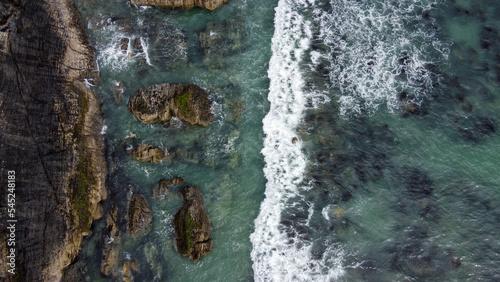 Waves break on the rocky shore of the Celtic Sea, top view. White foam on the waves. The coastline of the Atlantic. Turquoise sea water. Breathtaking seaside landscape. Aerial photo. View from above.