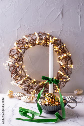 White burning candle, shiny glass cones, rustic wreath with  bright fairy ligts on grey  textured background. Place for text. Christmas postcad. photo