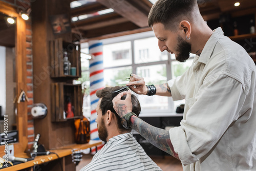 Bearded barber cutting hair of man in barbershop.