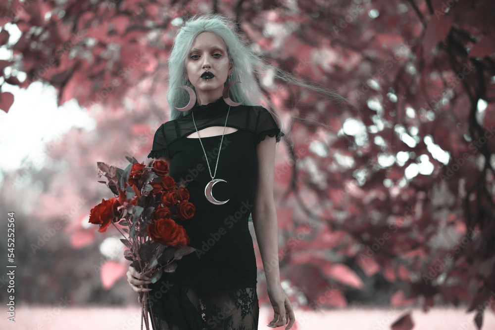 Obraz na plátně Dark goth girl standing in the forest, holding a bouquet of red roses, posing am