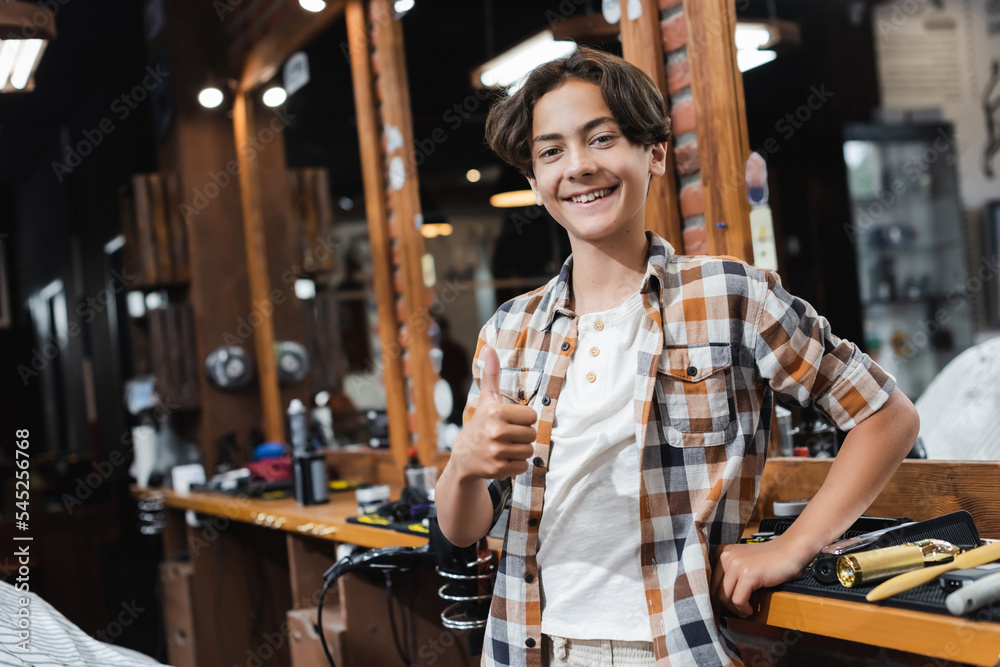Cheerful teenage boy showing thumb up near hairdressing tools and mirror in beauty salon. Cheerful teenage boy showing thumb up near hairdressing tools and mirror in beauty salon.