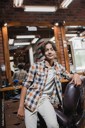 Positive teen boy looking at camera near armchair in blurred barbershop.