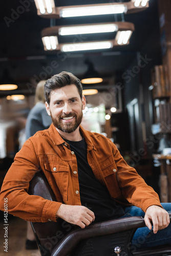 cheerful bearded man looking at camera while sitting in barbershop.