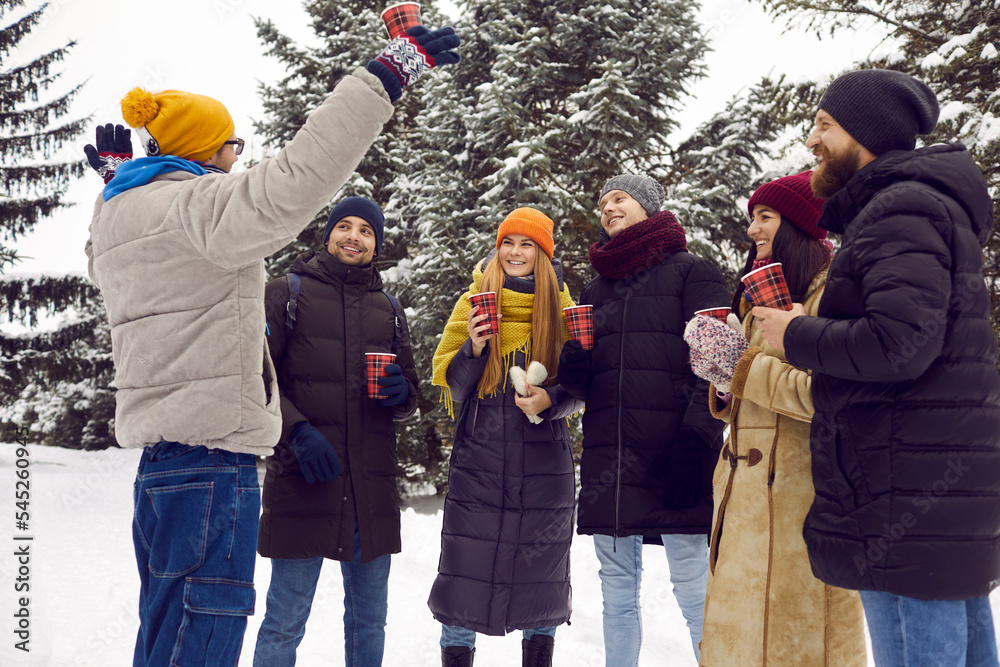 Group of happy cheerful people in puffers drinking coffee in fresh air among snow covered trees. Young male and female college or university friends in warm clothes having fun outdoors on winter break