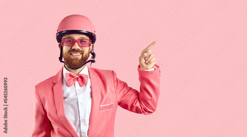Portrait of funny man with ginger beard and happy face expression wearing pink bike helmet, suit, bowtie and sunglasses standing isolated on pink background, smiling and pointing to side at copy space Portrait of funny man with ginger beard and happy face expression wearing pink bike helmet, suit, bowtie and sunglasses standing isolated on pink background, smiling and pointing to side at copy space