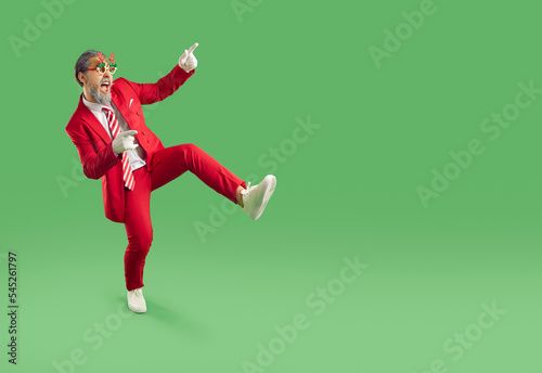 Happy excited goofy man in red suit and funny reindeer antler glasses standing on green background, pointing fingers to empty copy space side, advertising holiday sale or inviting to Christmas party
