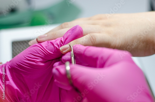 Female manicurist in pink gloves works with young hands and nails under a bright lamp. Process of cutting the cuticle with scissors