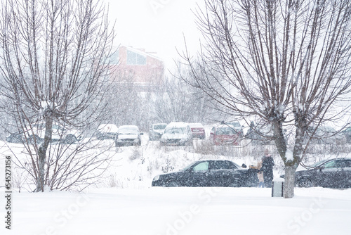 Winter city landscape. Parked cars, buildings and trees in the snow. Blizzard.