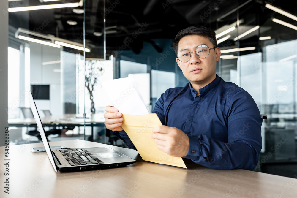 Young Asian male lawyer in glasses sitting in the office at a desk with a laptop. He holds an envelope with documents in his hands. He looks seriously into the camera. Young Asian male lawyer in glasses sitting in the office at a desk with a laptop. He holds an envelope with documents in his hands. He looks seriously into the camera.