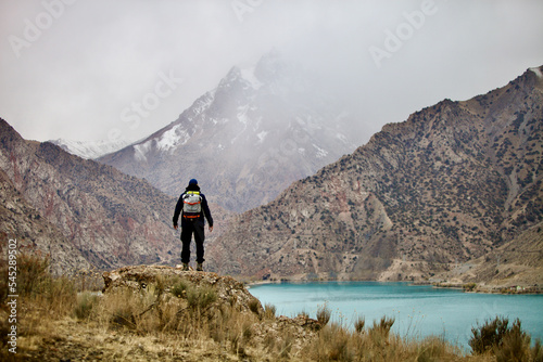 A man with a backpack stands on a mountainside and looks into the distance at a mountain lake and peaks. A lone hiker travels high in the mountains on a cloudy day. Independent hike.