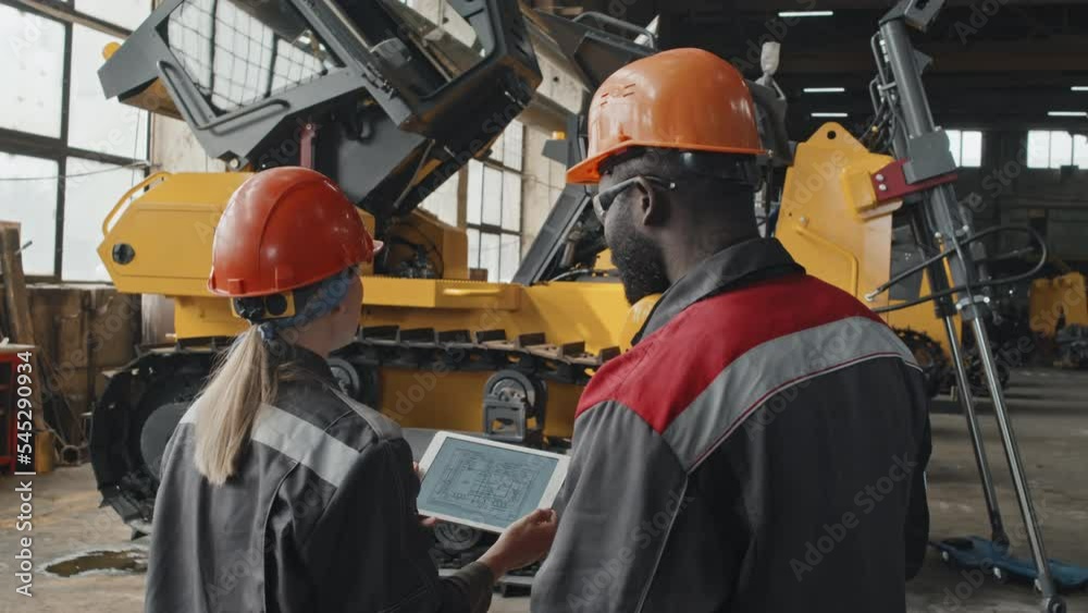 Rear view of two multiethnic engineers discussing tractor machine scheme on digital tablet while coworking at industrial plant Rear view of two multiethnic engineers discussing tractor machine scheme on digital tablet while coworking at industrial plant