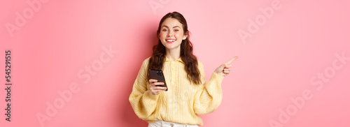 Beautiful modern girl holding smartphone and pointing finger aside, showing promotional text on copyspace while using mobile phone, standing over pink background