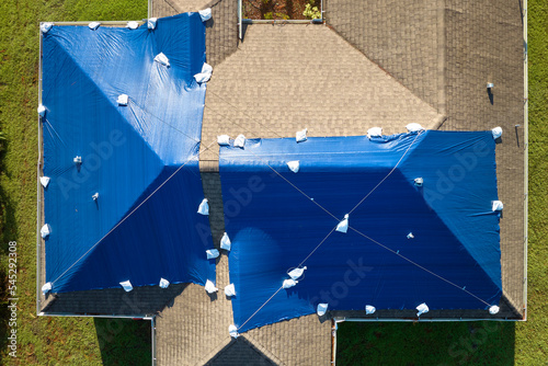 Aerial view of damaged in hurricane Ian house roof covered with blue protective tarp against rain water leaking until replacement of asphalt shingles photo