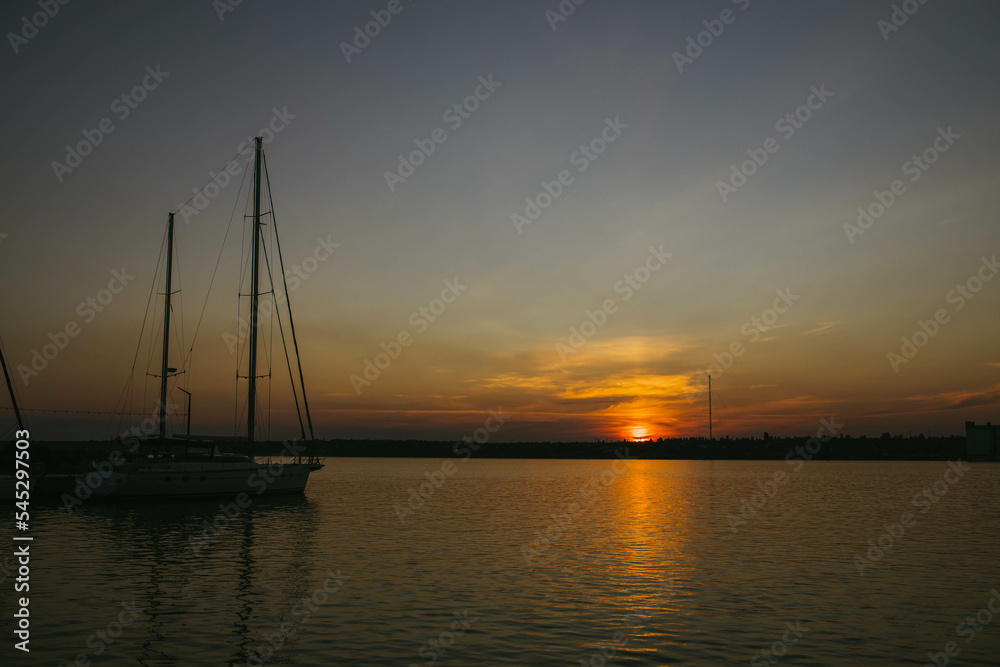 South Ukraine, Mykolaiv - August 21, 2021: Sunset in the river coast side, sun reflects into the water surface. Yachts and boats near the beach, people resting, Calm atmosphere in peaceful  city