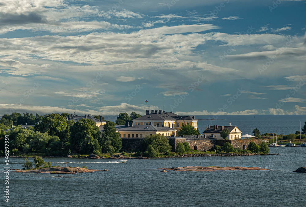 Helsinki, Finland - July 20, 2022: Suomenlinna Fortress from the sea. Ramparts and yellow buildings and terrain of the Naval Academy o Pikku Musta island under blue cloudscape. Islets in sea