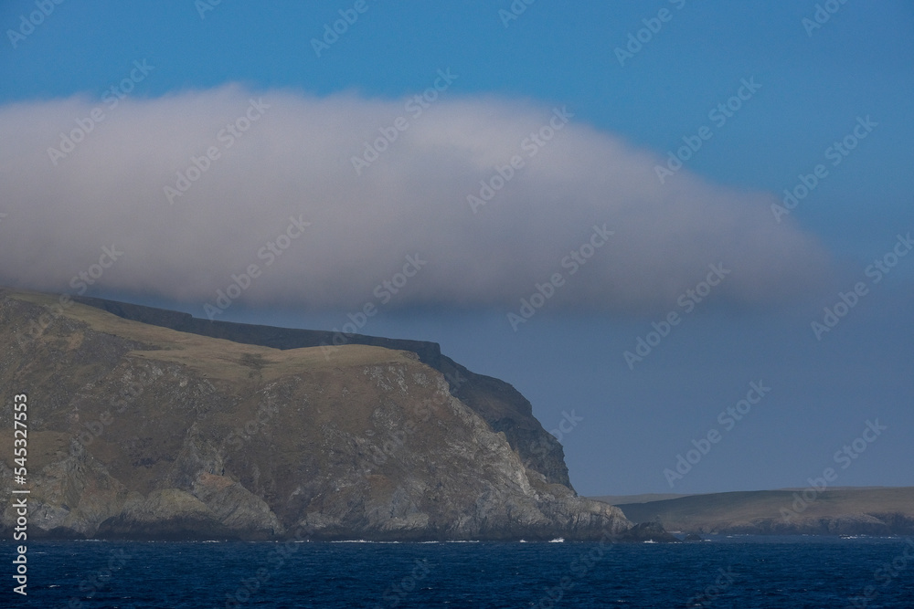 Cliffs and rocky coastline silhouette coast of Shetland Islands in Atlantic Ocean on sunny day with clouds, lighthouse and hills seen from cruiseship cruise ship liner with spray