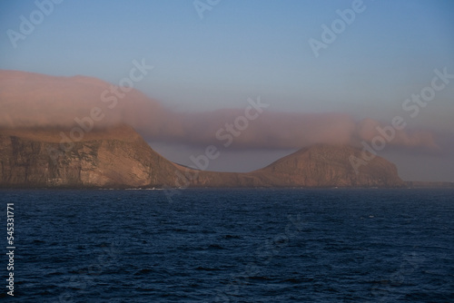 Cliffs and rocky coastline silhouette coast of Shetland Islands in Atlantic Ocean on sunny day with clouds  lighthouse and hills seen from cruiseship cruise ship liner with spray